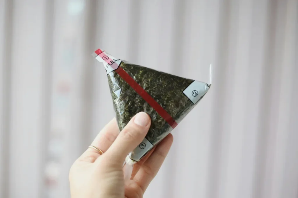 Hand holding a packaged Japanese onigiri rice ball wrapped in seaweed, a popular convenience store food in Japan.
